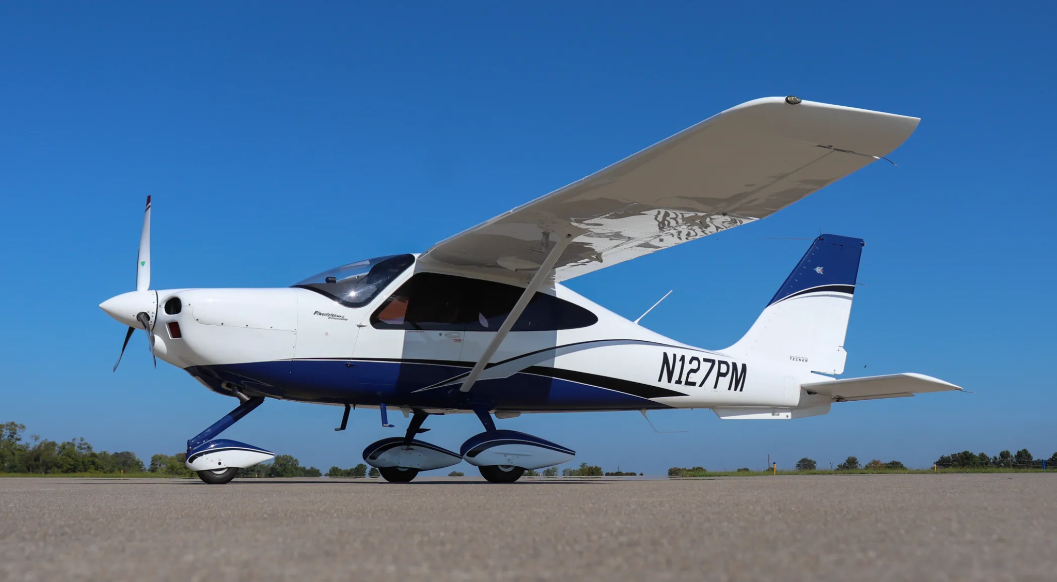 A side view of the white and blue Tecnam P2010 aircraft, registration N127PM, parked on an airfield.