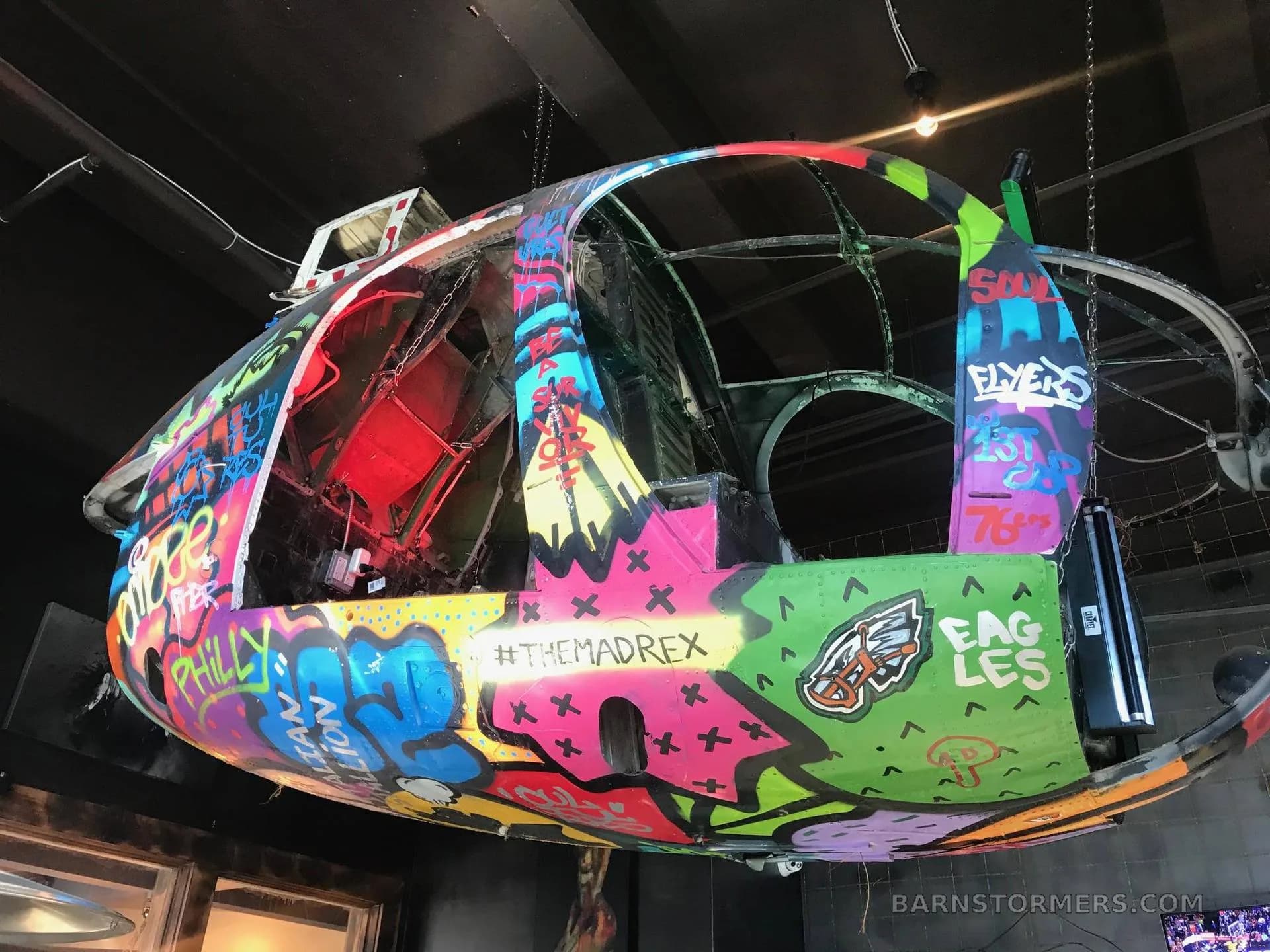 A colorful, graffiti-painted helicopter fuselage, possibly a Bell UH-1, hanging from a ceiling as a decorative art piece.