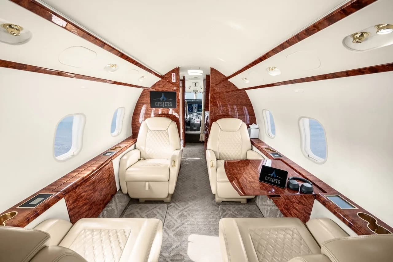 The forward section of a business jet cabin with two facing cream leather seats and a screen displaying 'CFSJETS'.