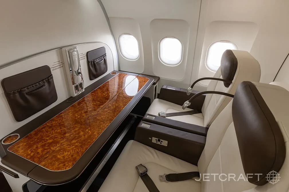 Two cream and brown leather aircraft seats facing a long, polished wooden table next to windows.