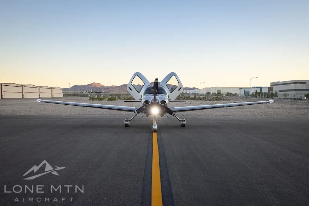 A silver Cirrus SR22T aircraft with registration N836ZJ is viewed head-on on an asphalt taxiway with its doors open and landing lights on, against a desert landscape.