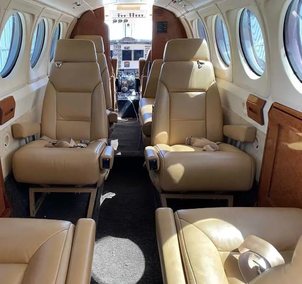 The aircraft cabin interior features tan leather seats arranged in rows, with the cockpit visible at the front.