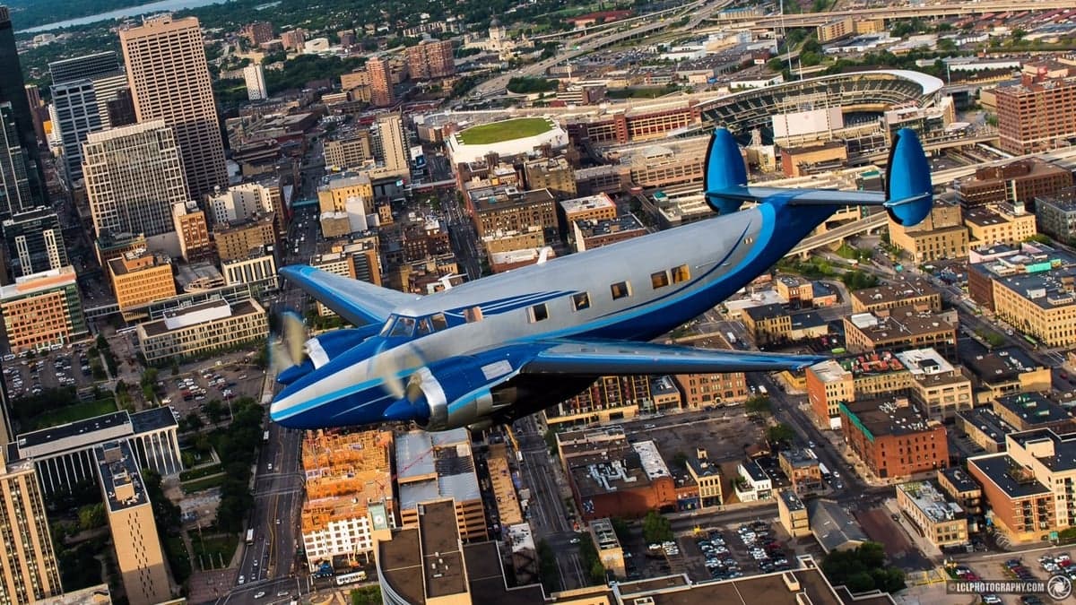 A blue and silver Beechcraft Model 18 flying over a city.