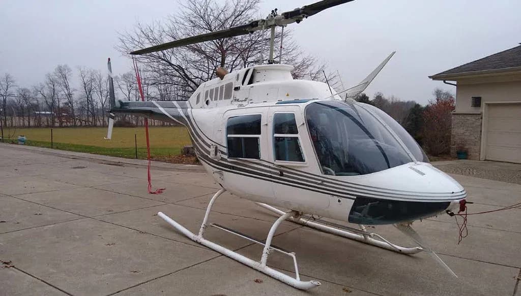 Front-side view of a white helicopter with dark grey stripes on a concrete driveway.