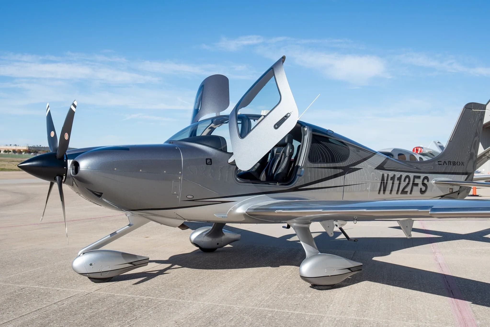A front-left view of a silver and dark grey Cirrus SR22T Carbon Edition aircraft (N112FS) with its gull-wing doors open on an airport tarmac.