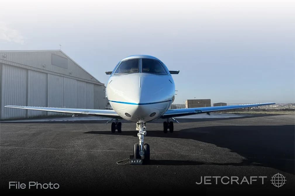 A white business jet with a blue stripe on the nose, seen from the front on an airfield with hangars in the background.