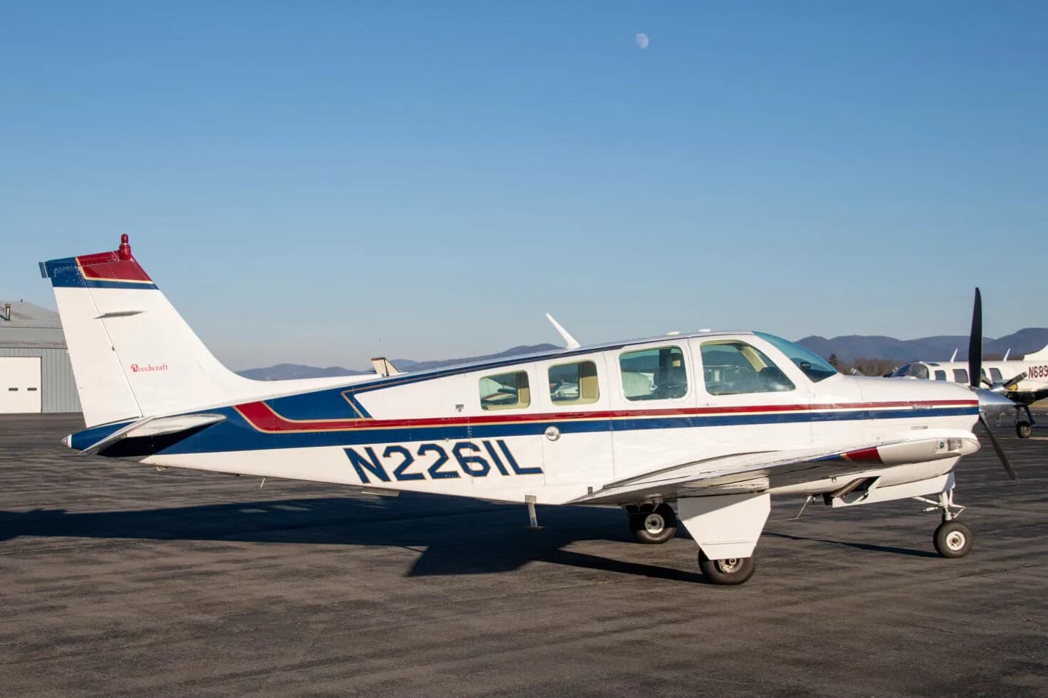 A white Beechcraft Bonanza N2261L with blue and red stripes parked on an airfield under a clear sky.