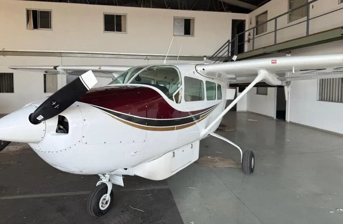 Three-quarter front view of a white and maroon single-engine propeller aircraft with a three-blade propeller, parked in a hangar.