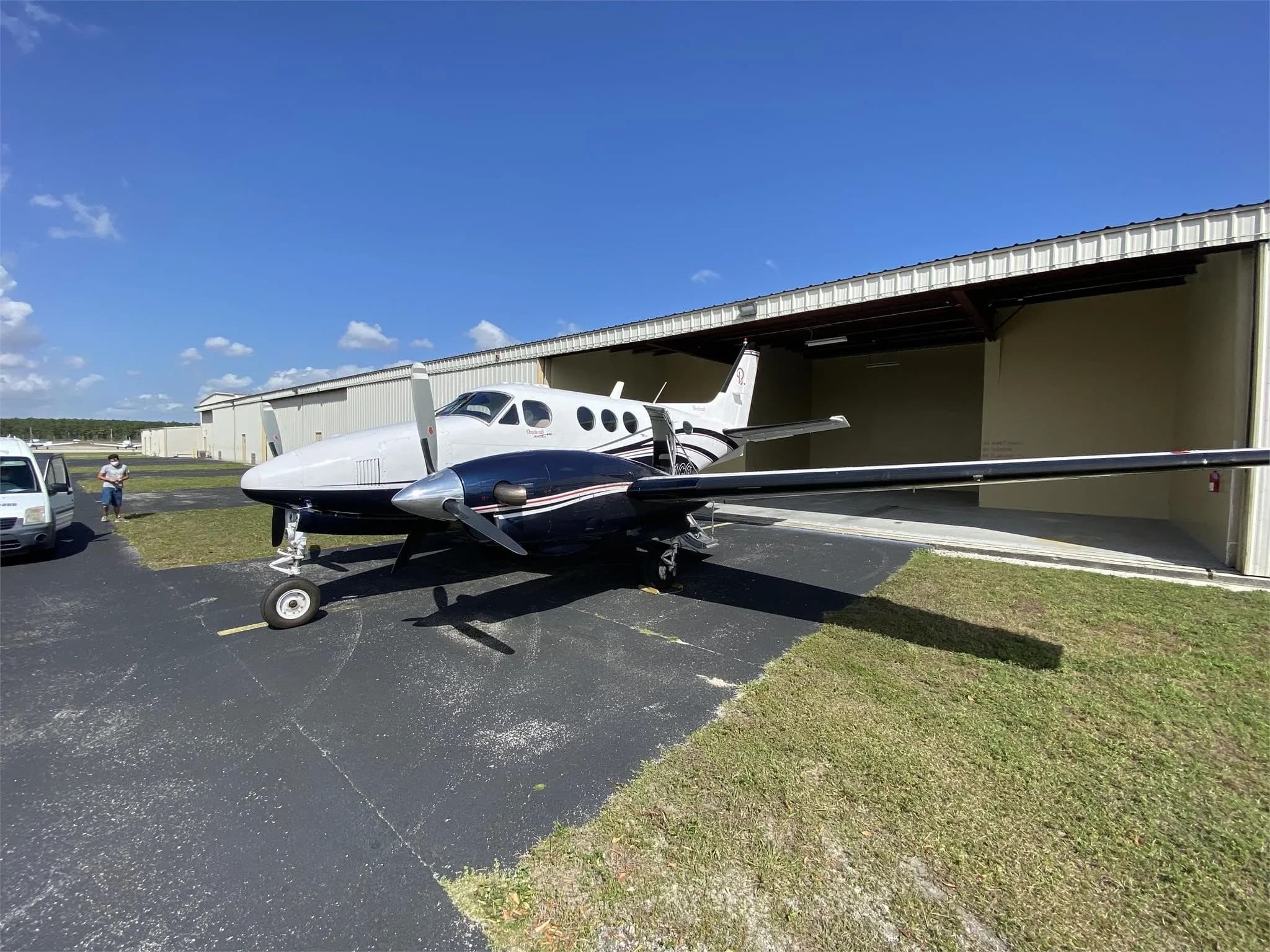 A white and dark blue Beechcraft King Air aircraft parked on an asphalt apron next to a hangar with a white van in the background.
