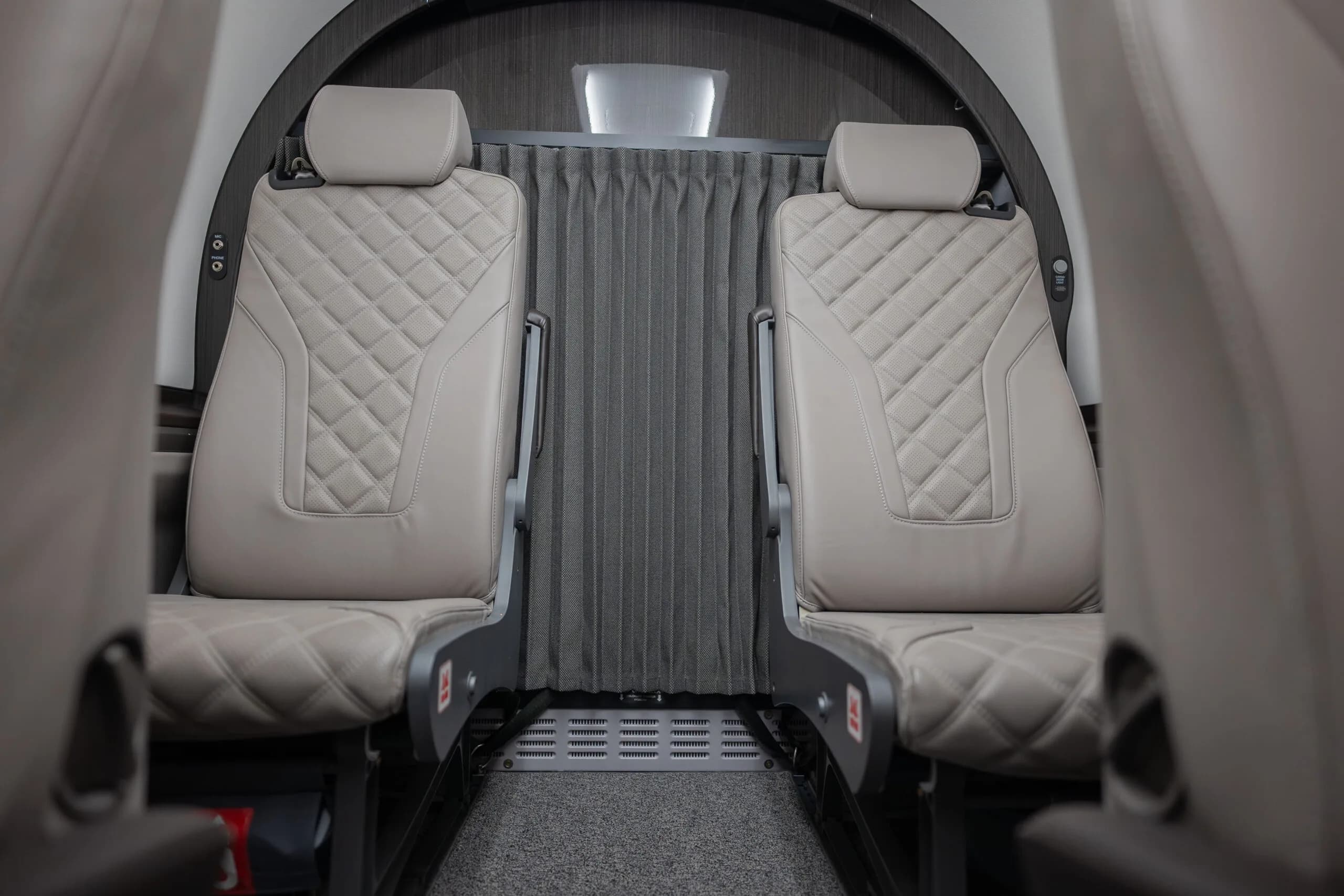 A close-up view of two light gray quilted leather seats in the cabin of a private aircraft, separated by a privacy curtain.