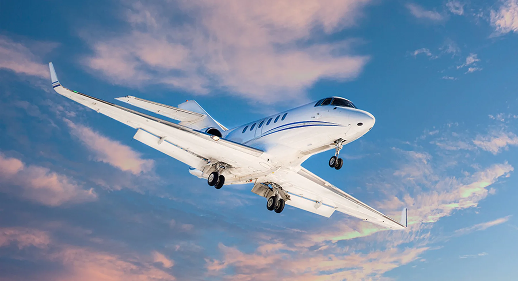 A white business jet with blue stripes flying with its landing gear extended against a blue and pink cloudy sky.