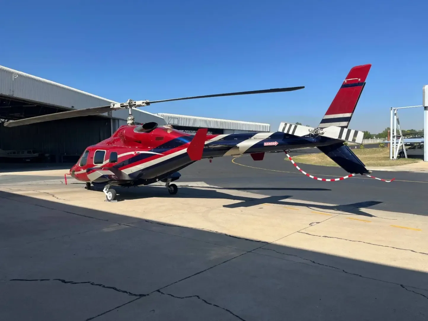 A red and white helicopter sits on the tarmac in front of a hangar.
