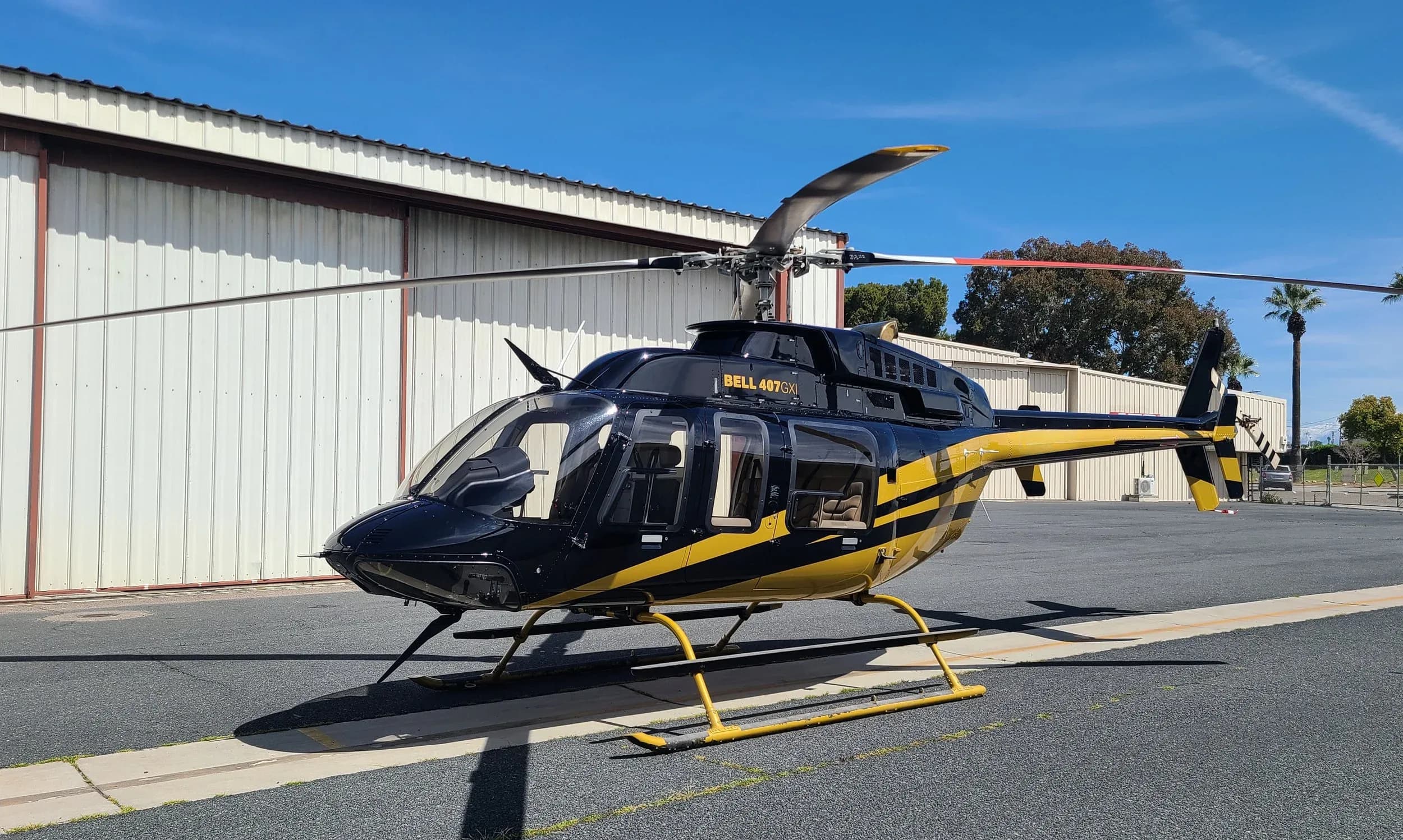 A black and gold Bell 407GX helicopter parked on an asphalt tarmac in front of a white hangar.