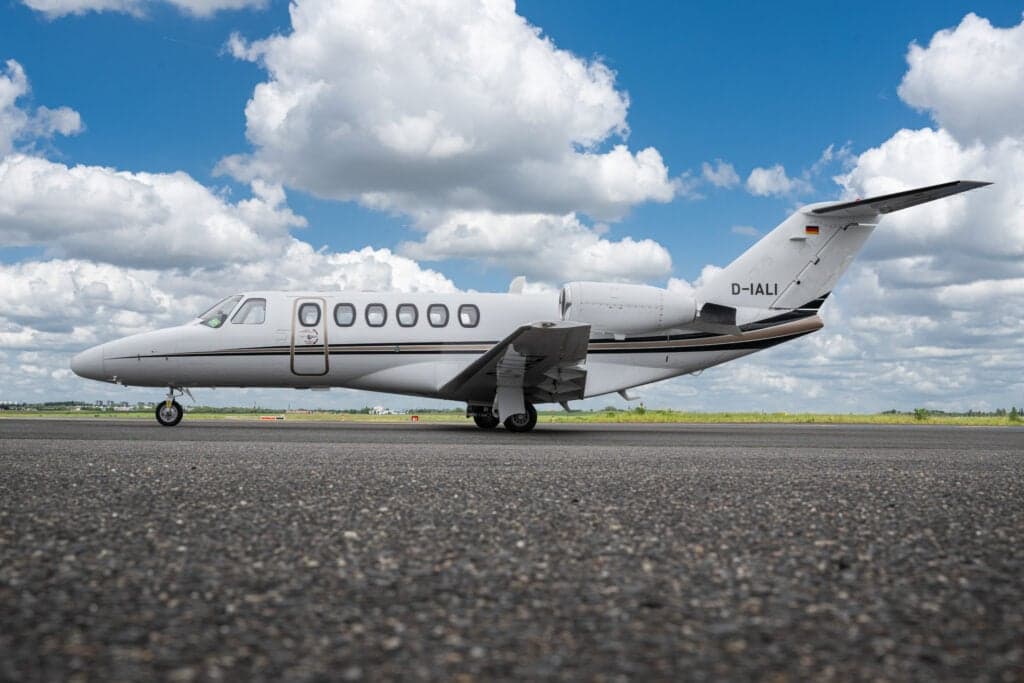 A white Cessna Citation CJ3 (D-IALI) private jet with black and gold stripes parked on an asphalt runway under a blue sky with white clouds, viewed from a low angle.
