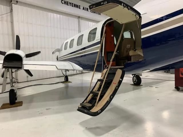 The side of a white and blue twin-engine turboprop aircraft with its passenger entry door and integrated stairs open in a hangar.