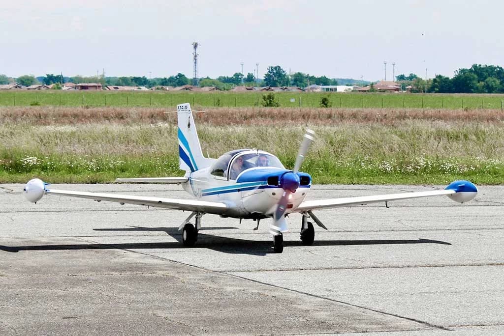 A white and blue single-engine aircraft with wingtip tanks is seen from the front on an airfield with grass in the background.