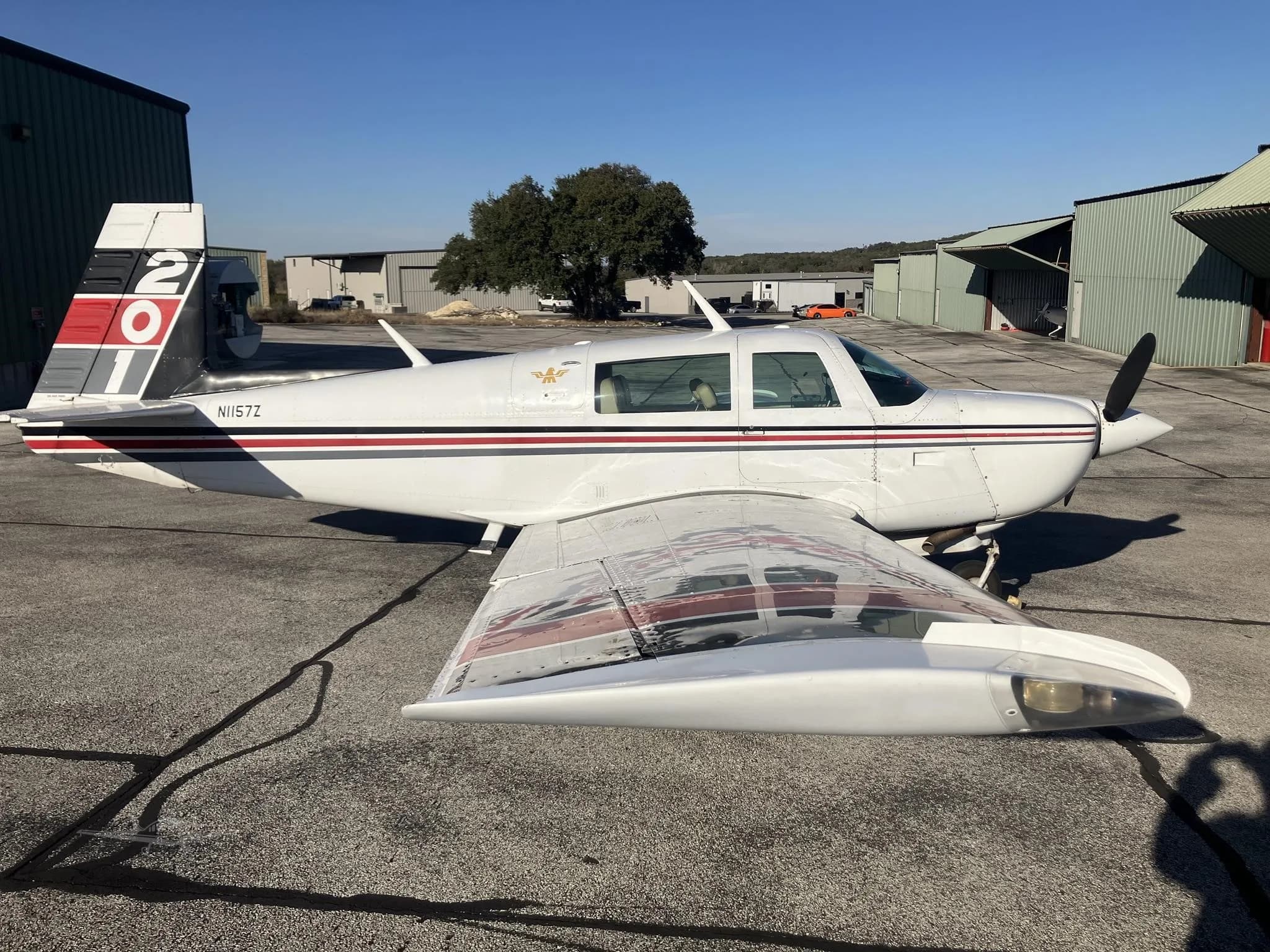 A white single-engine propeller aircraft with red and black stripes is parked on an airfield tarmac with hangars in the background.