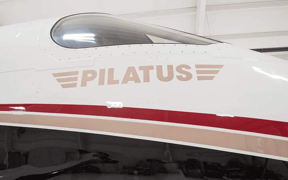 A close-up of the "PILATUS" logo in tan lettering with wing graphics on the white fuselage of an aircraft, above a red and tan stripe.