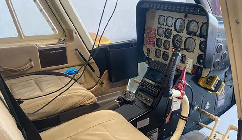 The cockpit of a helicopter showing the instrument panel, controls, and tan pilot's seat.