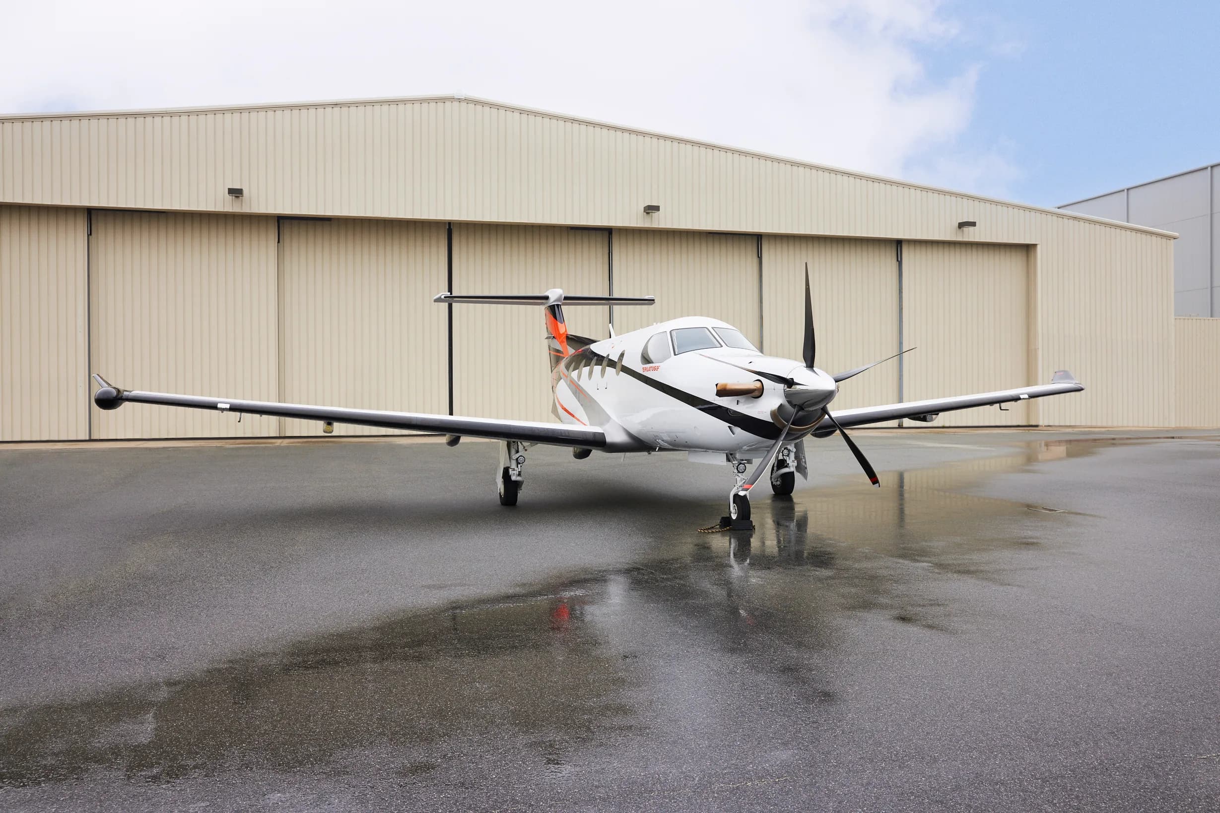 A white Pilatus PC-12 aircraft with black and orange accents parked on a wet tarmac in front of a hangar.