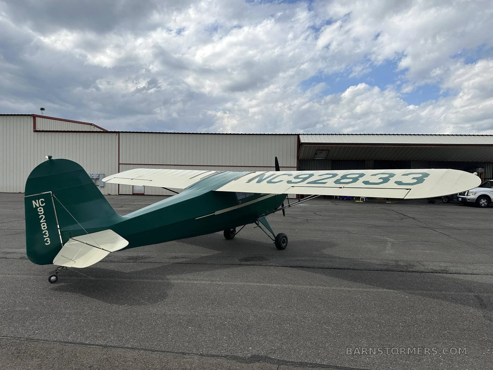 A green and cream vintage aircraft with tail number NC92833 is parked on an asphalt tarmac in front of a hangar under a cloudy sky.