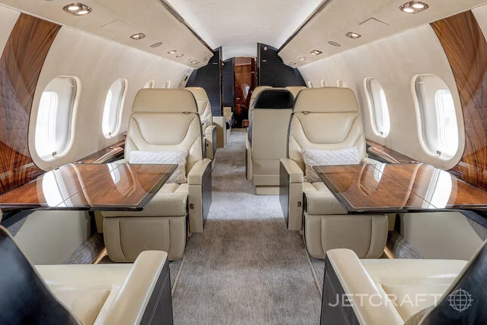 The elegant cabin interior of a Bombardier Global business jet, featuring beige leather seats with pull-out tables and wood accents.