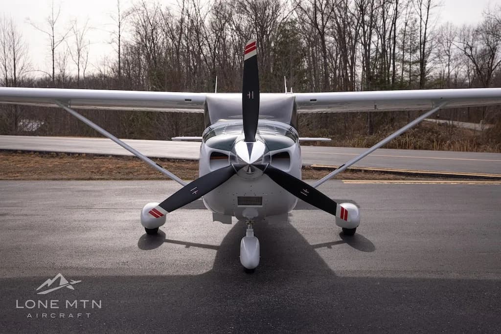 A head-on view of a white and dark green Cessna Skylane N915LS with a three-bladed propeller, parked on an asphalt ramp.