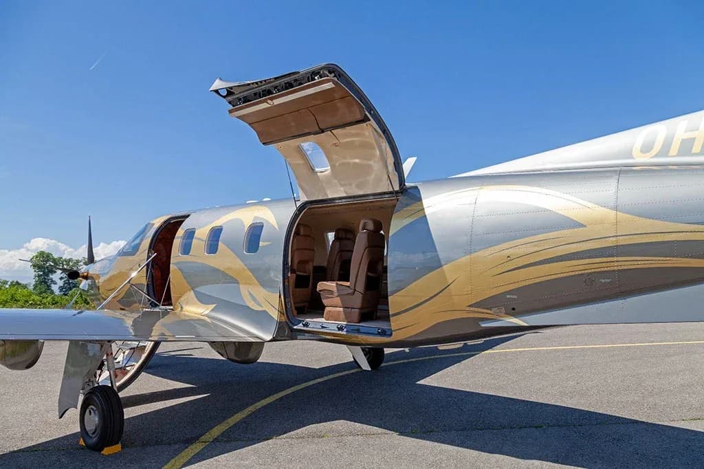 A side view of a silver and gold Pilatus PC-12 aircraft with its large cargo door and passenger door open, revealing the cabin interior.