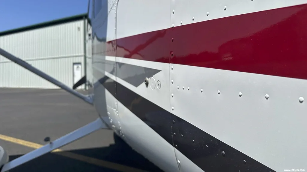 A close-up view of the white fuselage of a Cessna 182, showing red and gray stripe details and rivets.