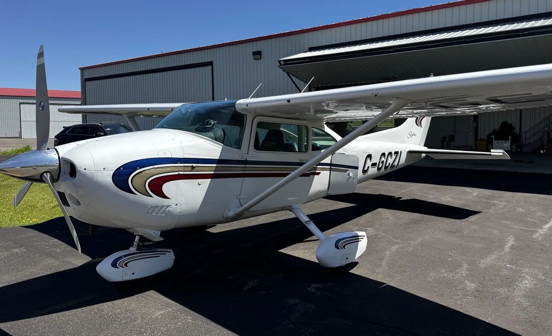 A white Cessna Skylane C-GCZI with blue, gold, and red stripes, parked on an asphalt ramp in front of a hangar.