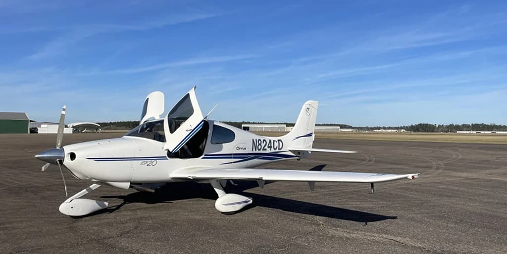 A white Cirrus SR20 aircraft with blue accents, registration N824CD, parked on an asphalt runway with its doors open, under a clear blue sky.