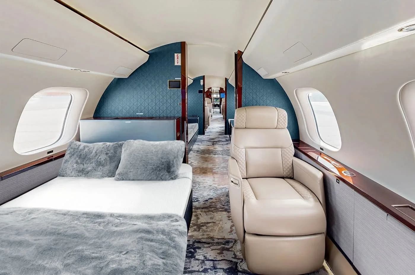 A private jet cabin featuring a bed with grey bedding on one side and a single light beige quilted leather seat on the other, with a blue patterned wall in the background.