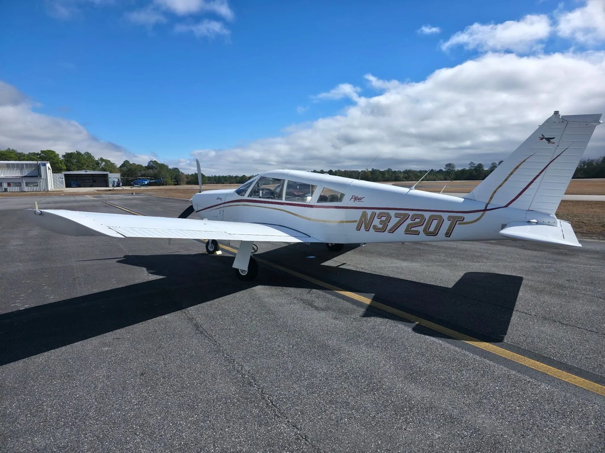 A three-quarter rear view of the white Piper Arrow N3720T parked on an asphalt apron under a blue sky with scattered clouds.