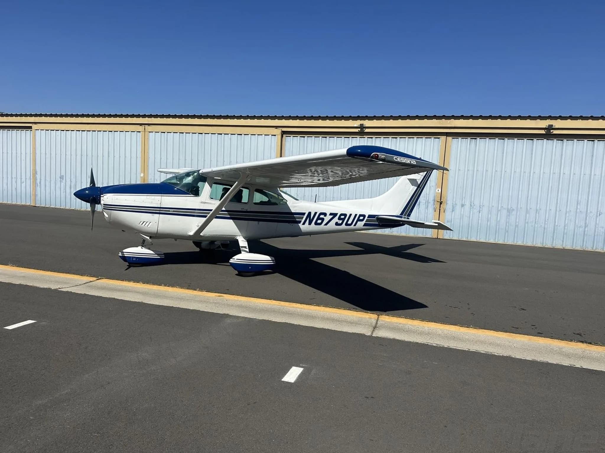 A white and blue Cessna 182 Skylane aircraft parked on an asphalt ramp next to hangars.