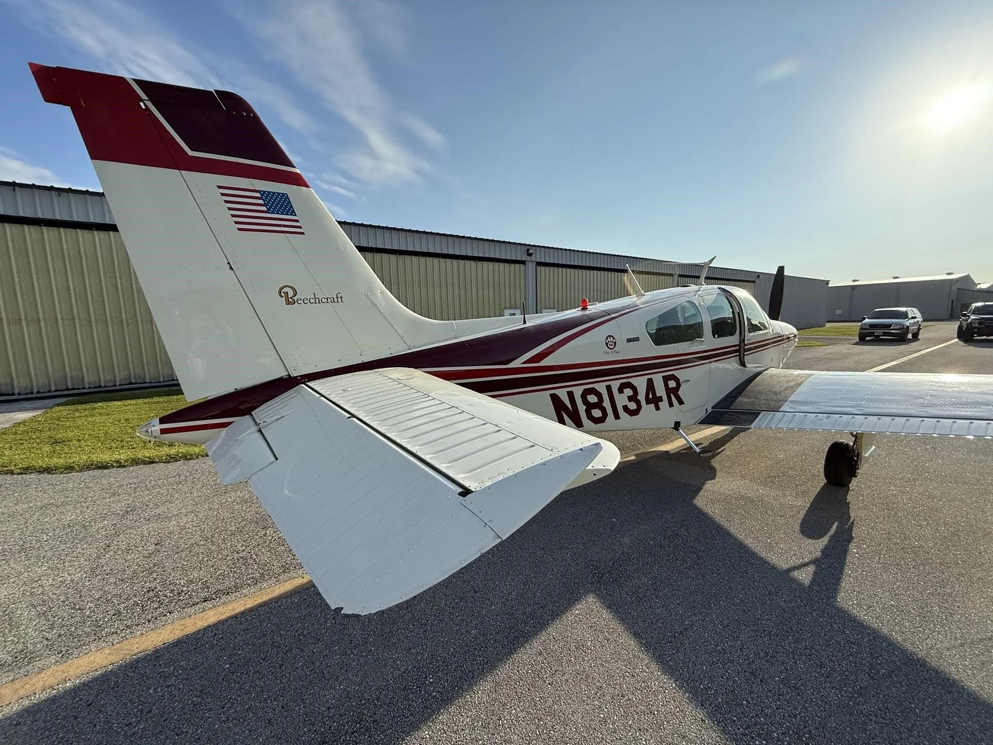 A white Beechcraft aircraft with maroon stripes, tail number N8134R, is parked on an asphalt ramp, viewed from the rear with hangars in the background.