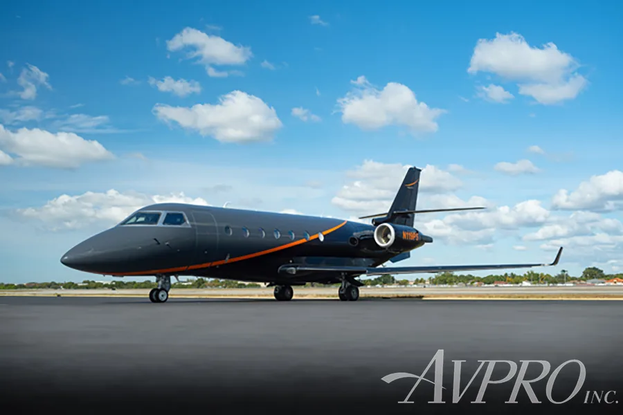 A Gulfstream G200 business jet, registration N119PS, with a dark gray and black paint scheme with orange accents, parked on an airfield under a blue sky.