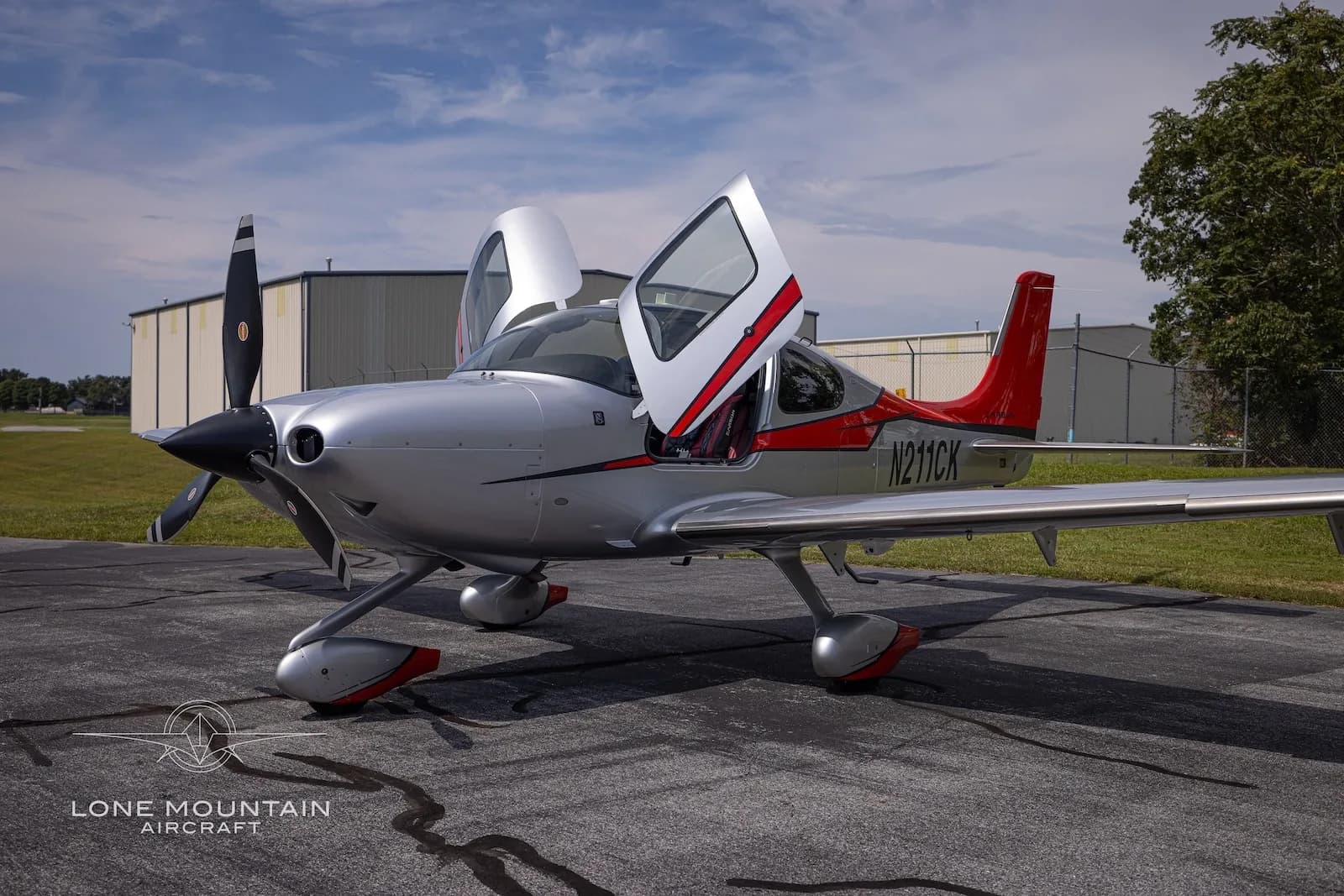 A silver and red Cirrus SR22T aircraft with registration N211CK is parked on an asphalt ramp with its gull-wing doors open.