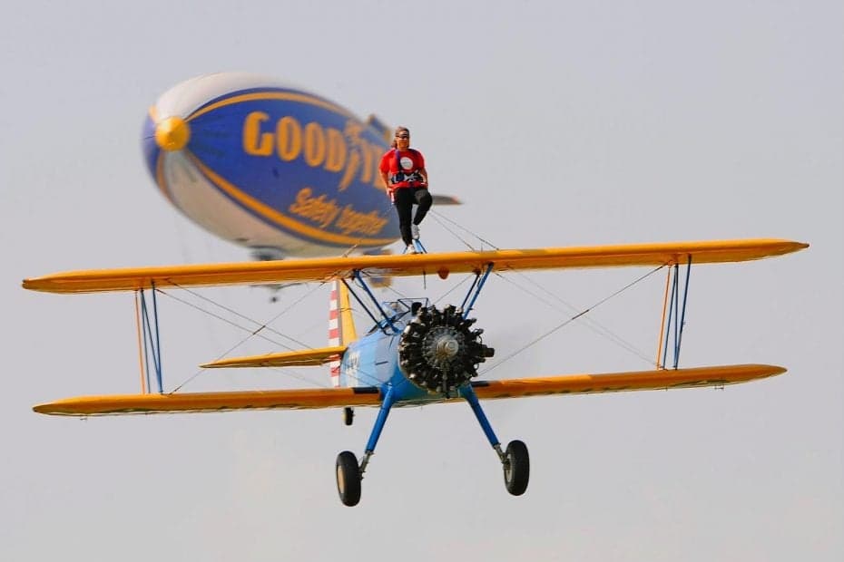 A Stearman biplane with a wing walker standing on top, with a Goodyear blimp in the background.