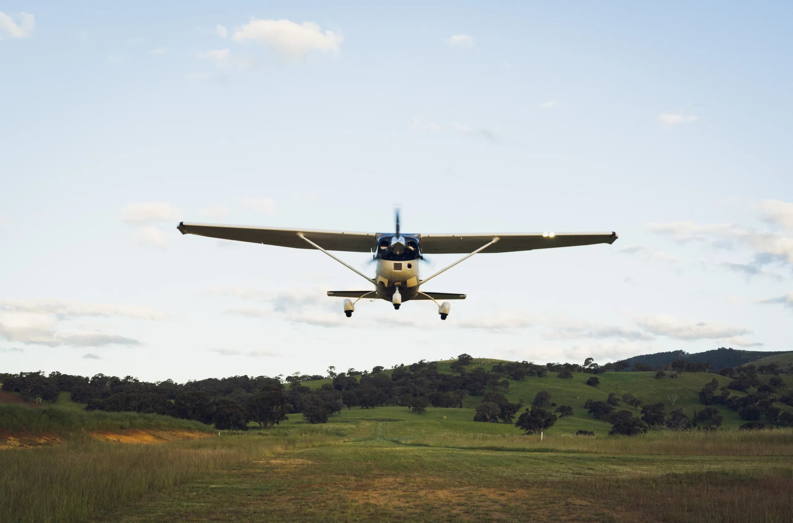A white single-engine high-wing aircraft with blue accents flying low over a grassy airstrip with rolling hills in the background.