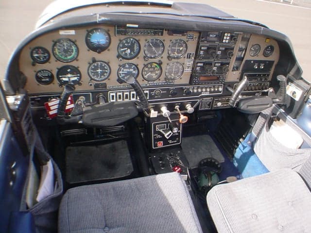 The cockpit of a Grumman AA-5 Traveler aircraft, showing the instrument panel, controls, and front seats.