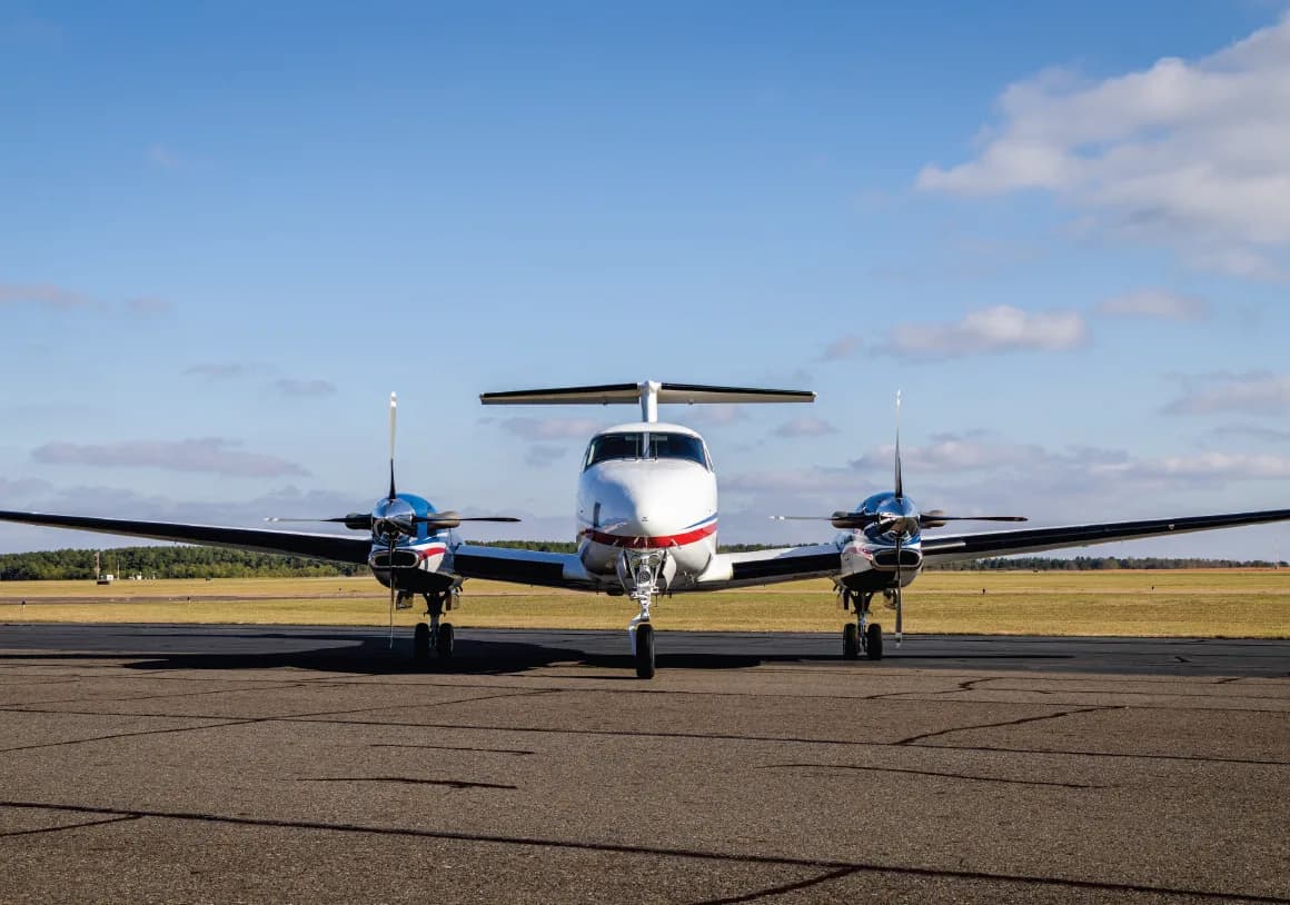 Front view of a white Beechcraft King Air 360 with blue and red stripes on a tarmac under a blue sky.