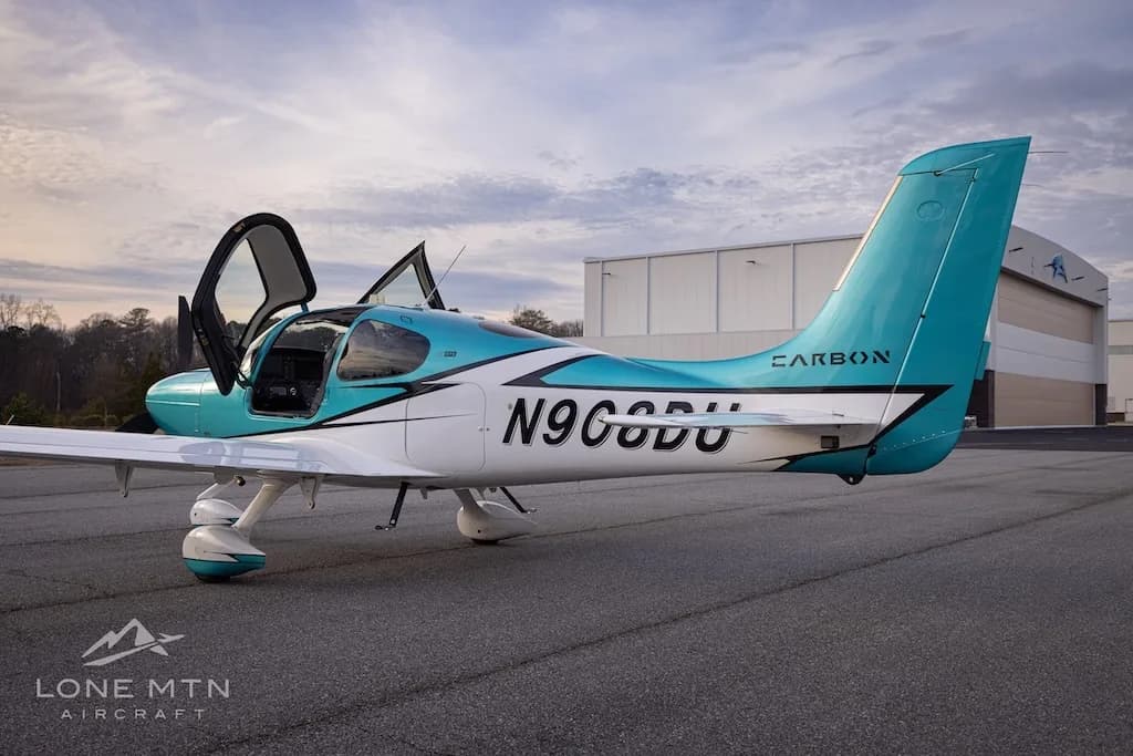 A rear-side view of the teal and white Cirrus SR22 aircraft, N908DU, with its doors open, parked on an airfield.