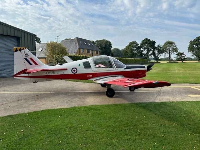 A red and white Bulldog aircraft parked on a paved area next to a building and grassy field.