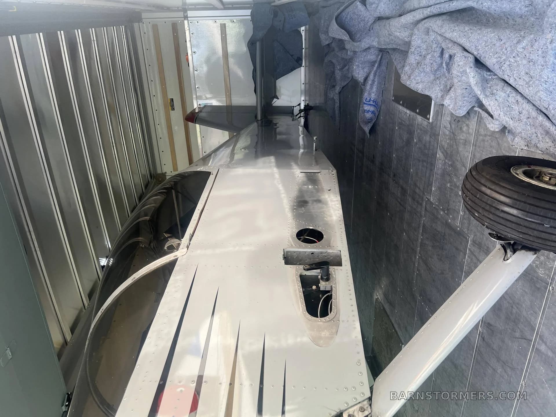 A silver aircraft fuselage with black stripes and a landing gear wheel, partially disassembled inside a trailer.
