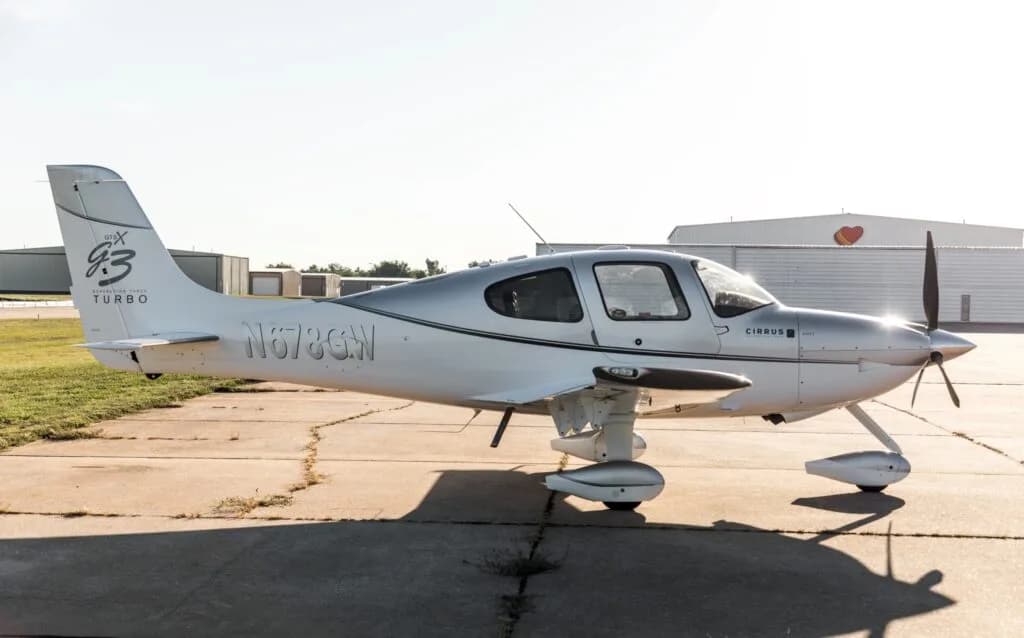 A side view of a silver and white Cirrus SR22 G3 Turbo aircraft (N678GW) on an airfield under bright sunlight.