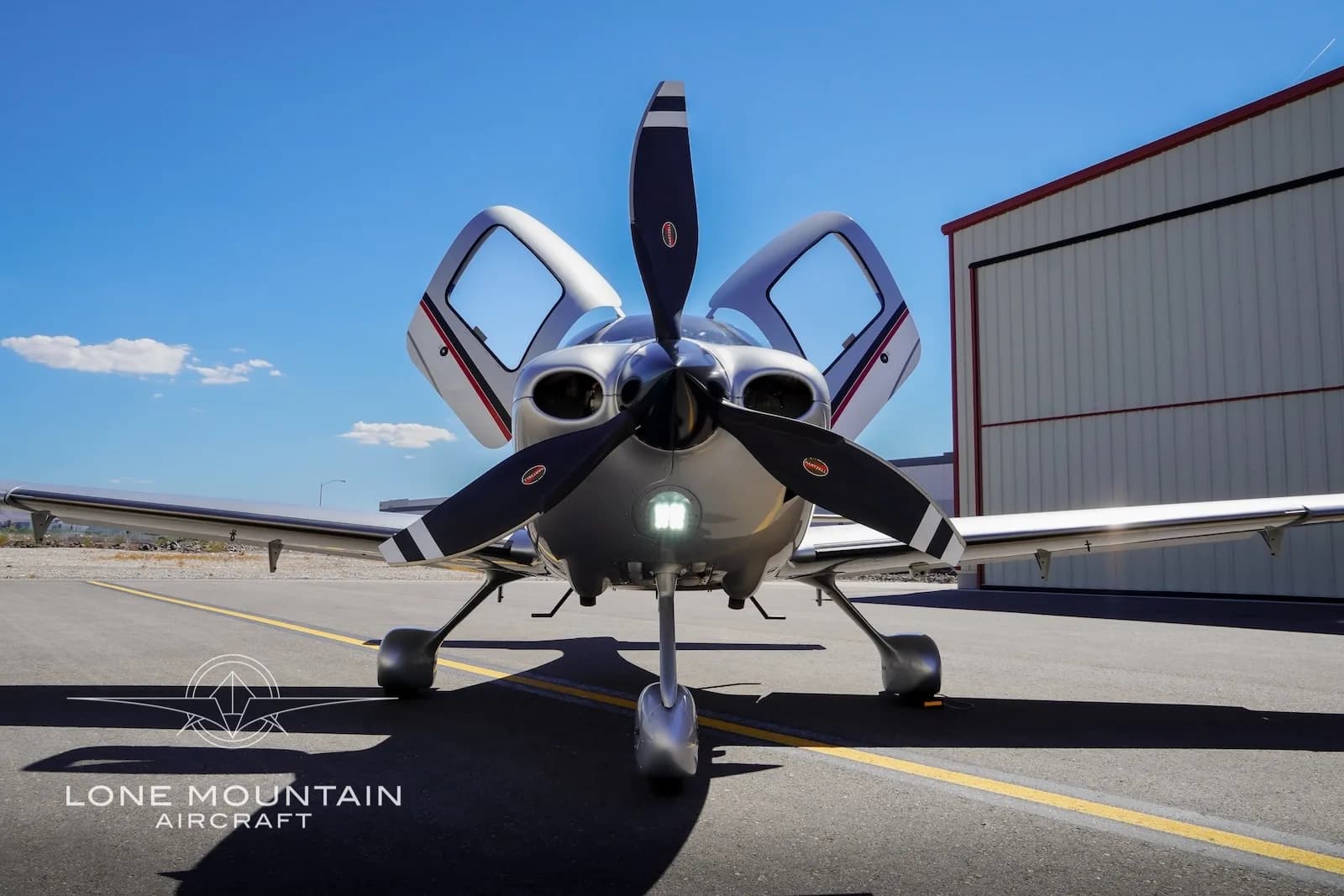 A head-on view of a silver Cirrus SR22 aircraft, registration N677RC, with its propeller blades vertical and both gull-wing doors open.