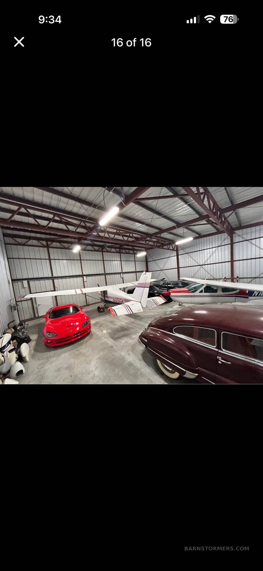 A white single-engine aircraft with red stripes, a red sports car, and a classic car are parked inside a metal hangar.