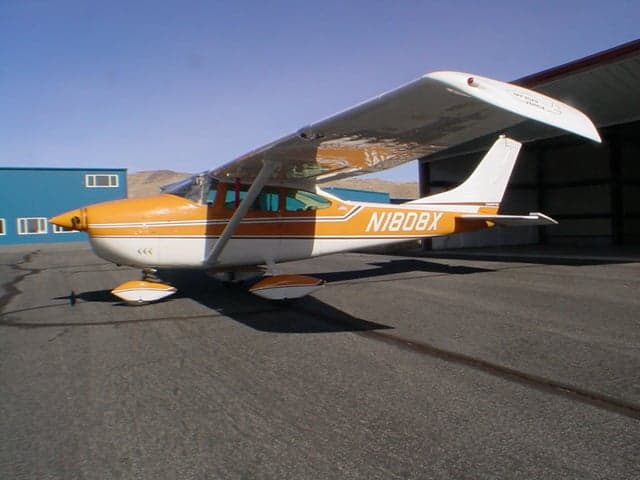 A Cessna 172 aircraft with an orange and white paint scheme, registration N1808X, parked on an asphalt ramp next to a blue hangar.