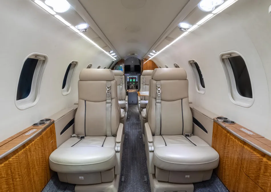 The interior of a business jet cabin, featuring beige leather seats and wood paneling along the windows.