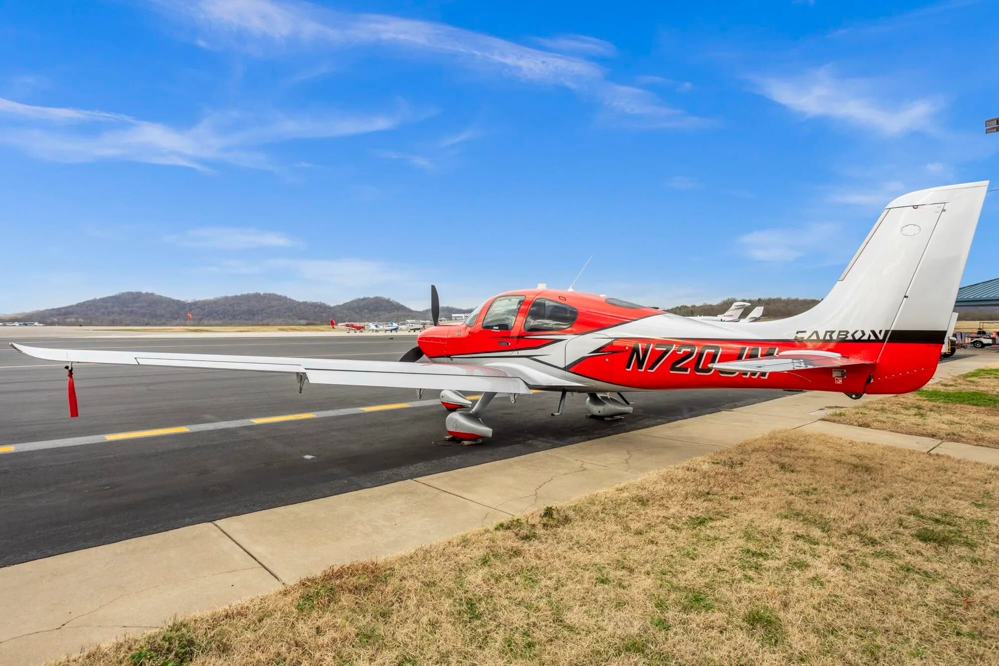 A side view of a red and silver Cirrus SR22 aircraft with registration N720JM parked on an airport tarmac with mountains in the background.
